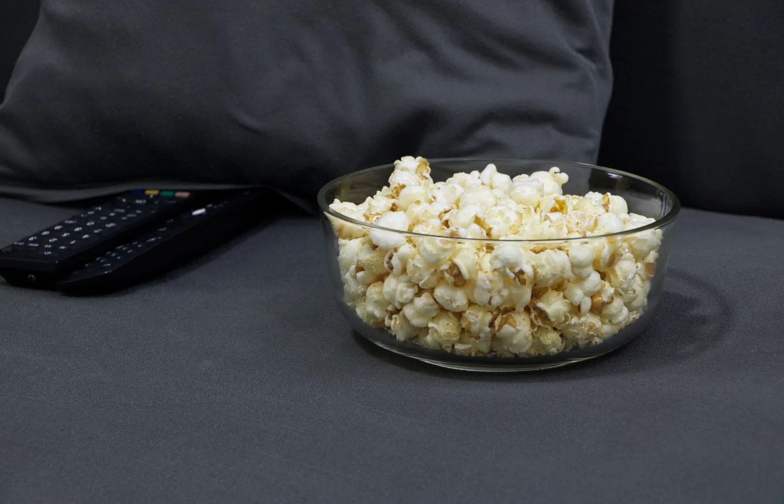 Glass bowl of popcorn beside TV remotes on a gray sofa