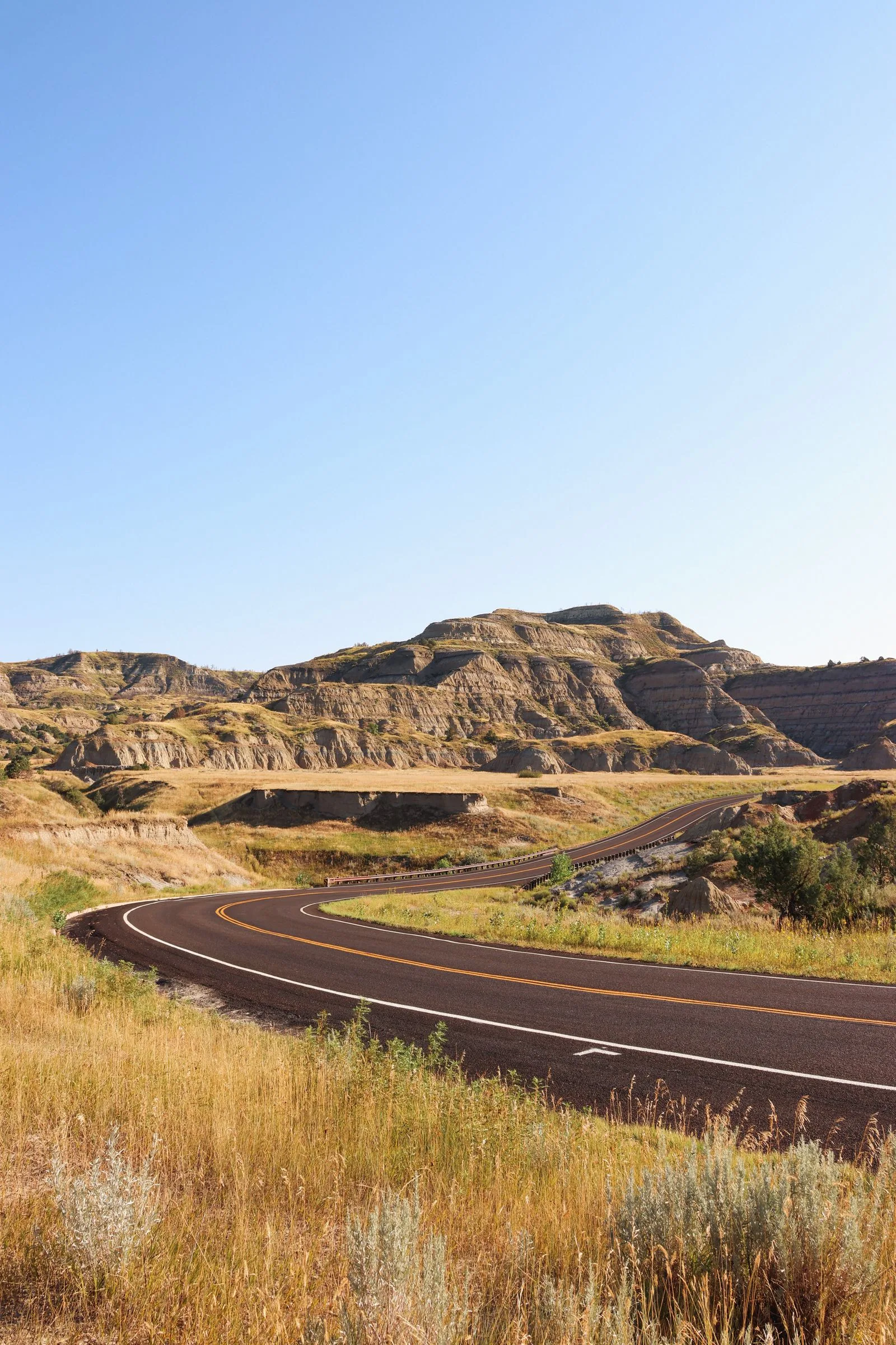 Two-lane road curving through dry, rolling hills under a blue sky